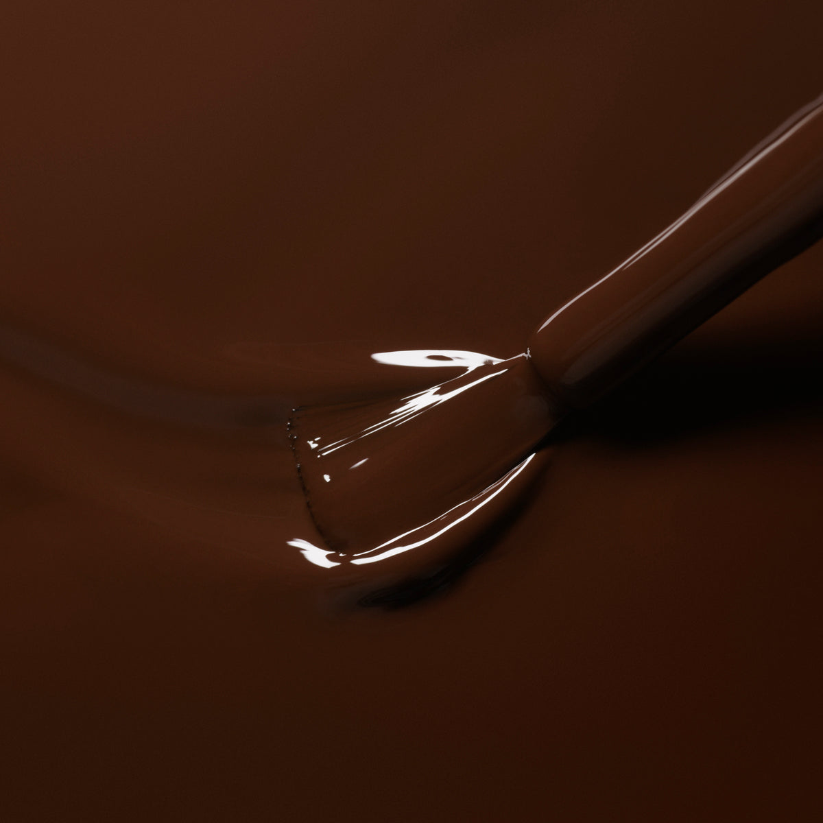 A close-up of a brush applying Manucurist’s Clove nail polish, with bristles partly submerged and the chocolate brown shade reflecting light in a glossy finish.