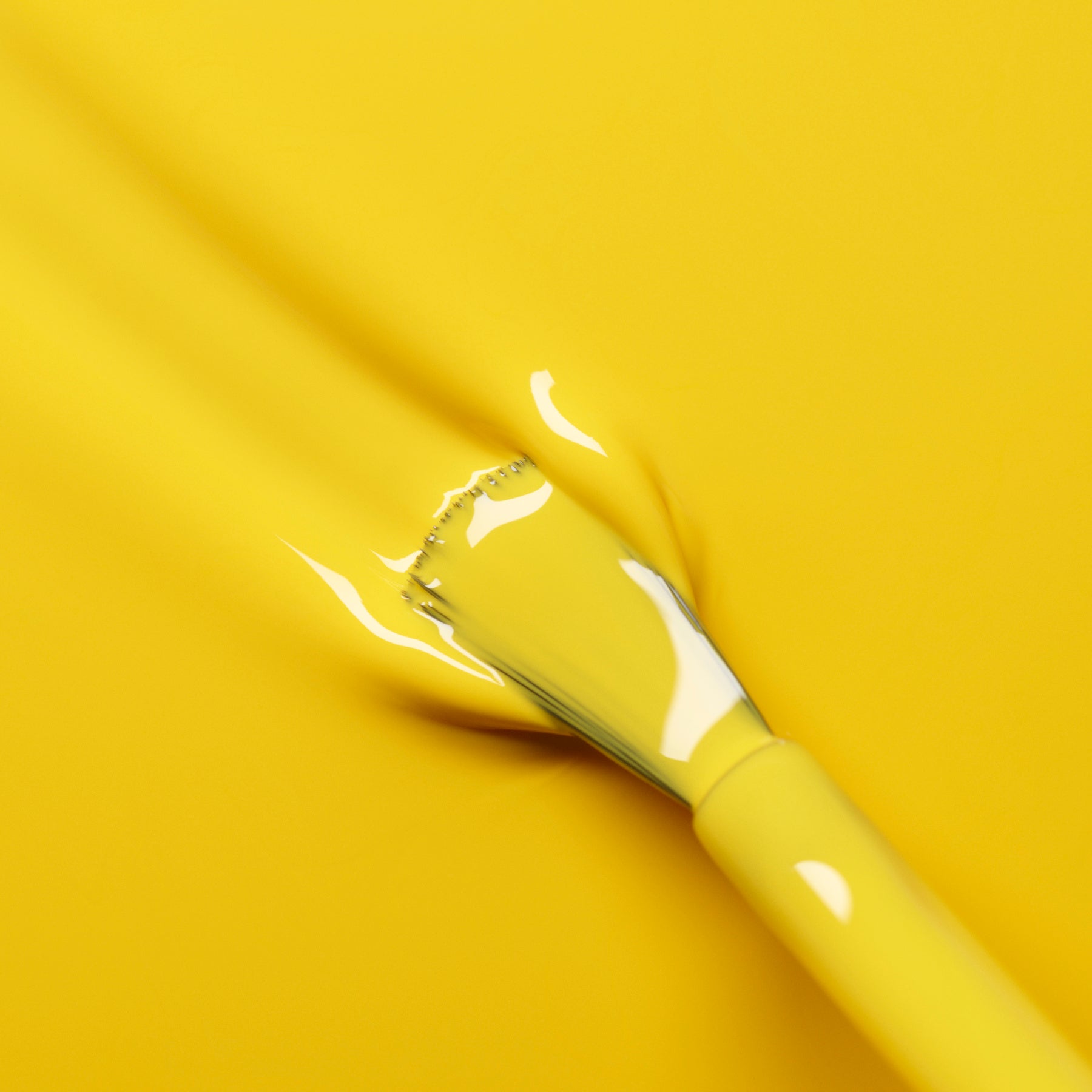 A Manucurist Gold Button brush rests on a smooth, vivid yellow surface, blending in as it leaves bold strokes of matching color.