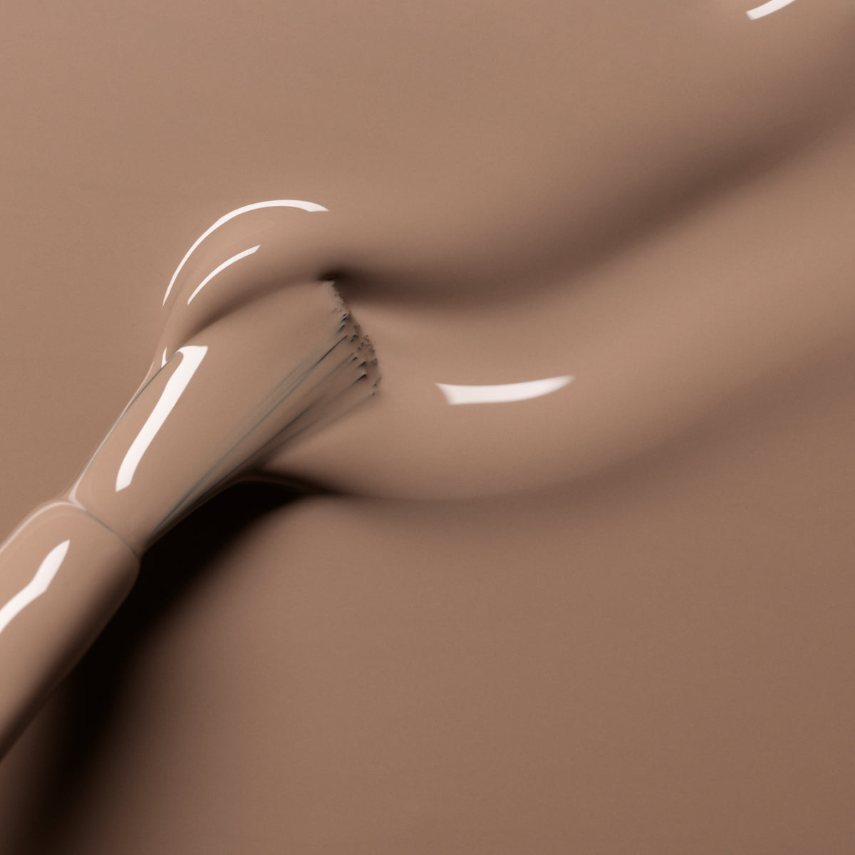 A close-up of a makeup brush spreading smooth, beige foundation evokes the glossy curves and creamy finish of Manucurist’s Orme nail polish, reflecting its chic café au lait tones and luminous allure.