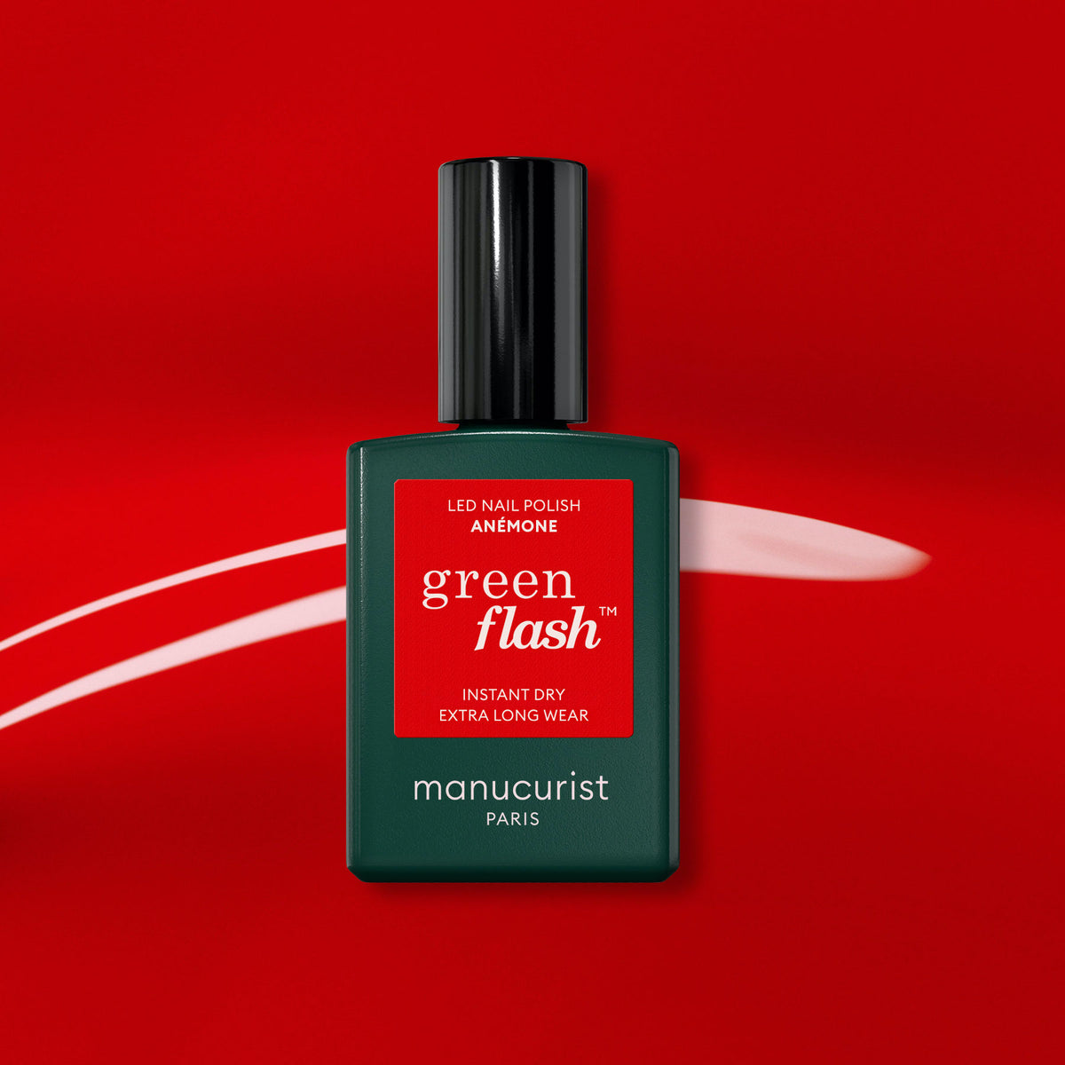 A bottle of Manucurist Anémone Green Flash gel polish stands upright on a red background with a white brushstroke behind it. The dark green bottle, black cap, and red label emphasize its rich, juicy red shade.
