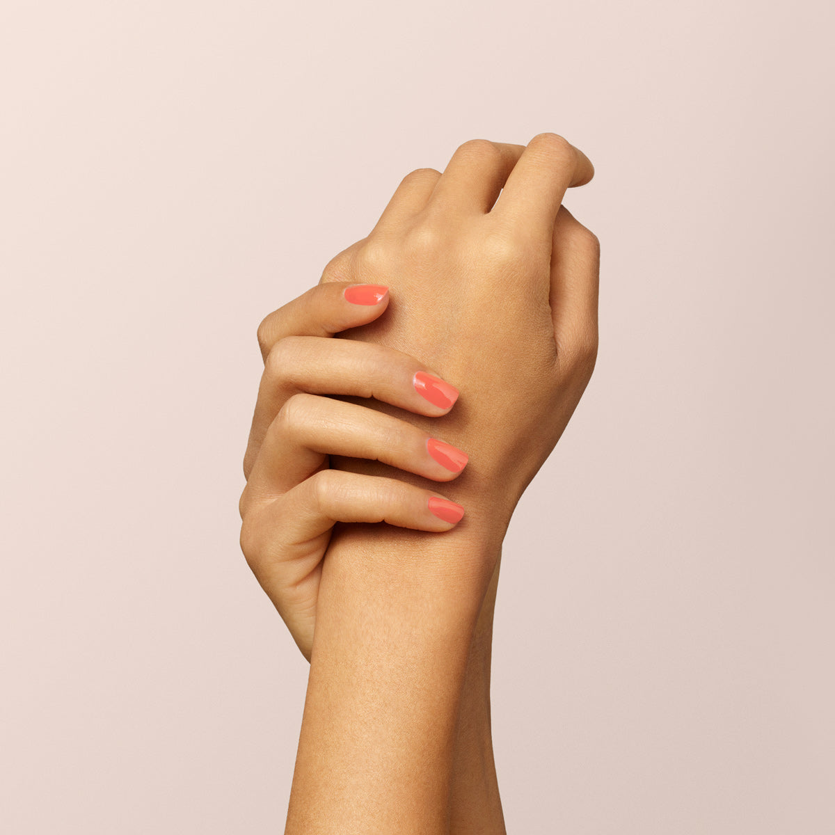 A close-up of two hands gently clasped, displaying neatly manicured nails painted in Manucurist’s Bird of Paradise, a glossy coral orange gel polish, set against a plain light beige background.