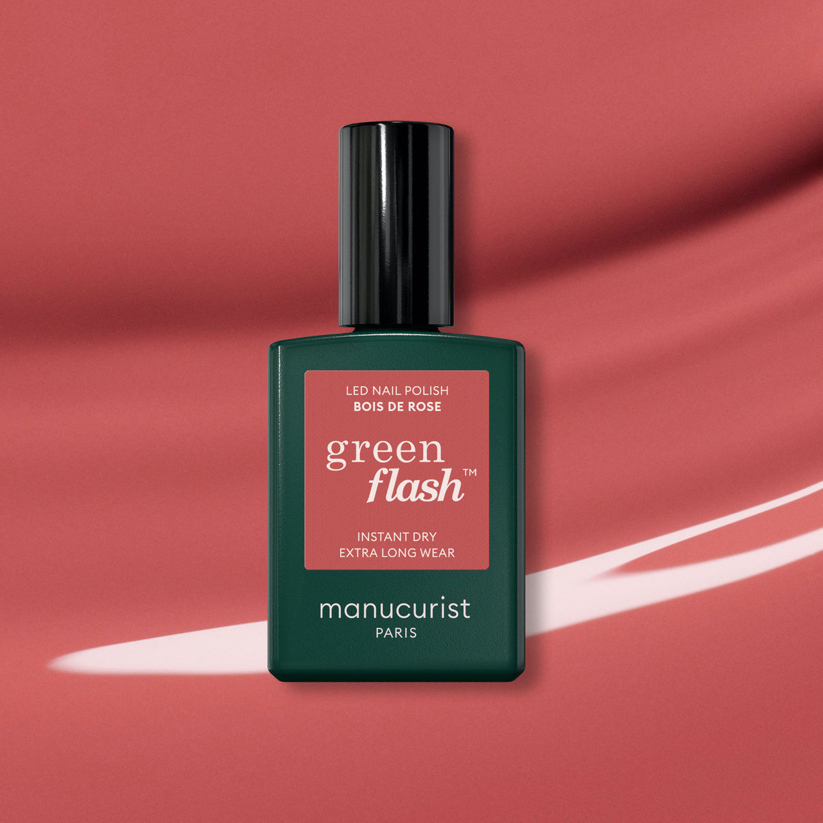 A bottle of Manucurist Bois de rose polish, a rosewood pink shade, is displayed on a pink background featuring a white brushstroke design.