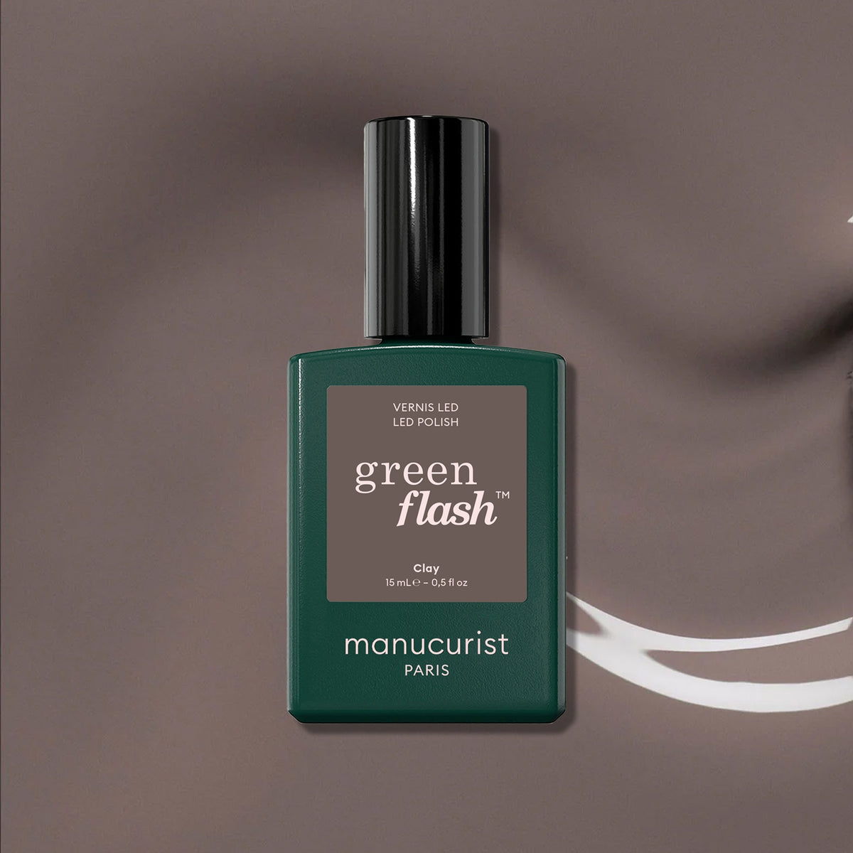 A bottle of Manucurist Green Flash gel polish in the shade Clay stands upright on a brown background, featuring a dark green bottle with a rectangular label—ideal for lovers of taupe gel polish.