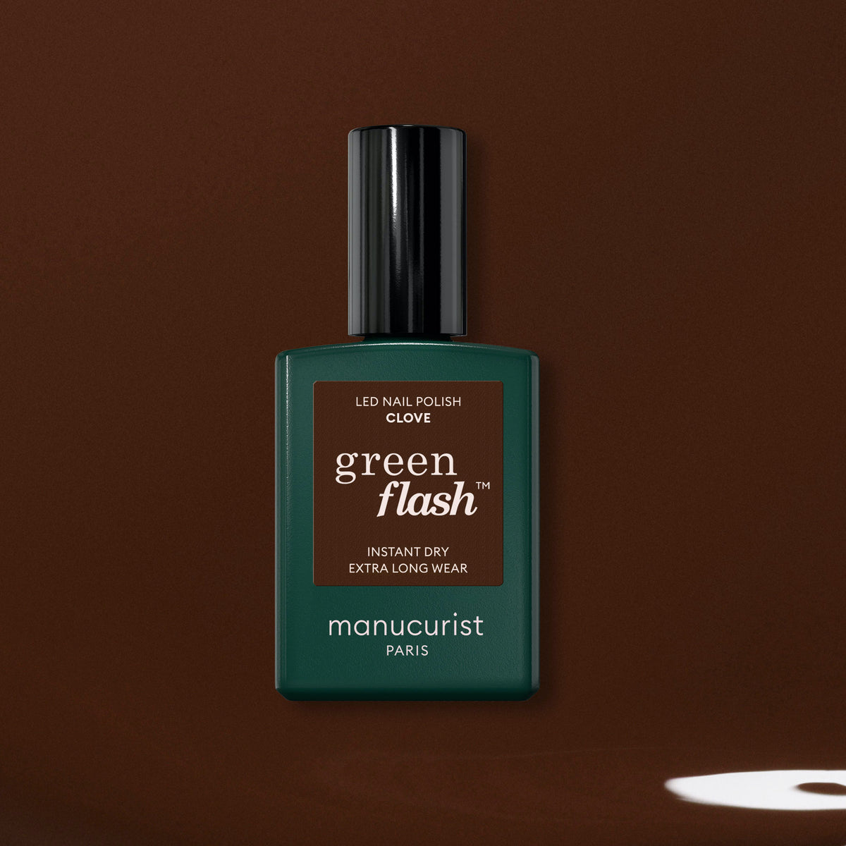 A bottle of Manucurist Clove Green Flash gel polish, with a green and brown label, is shown against a dark chocolate brown background featuring a small white spot in the lower right corner.