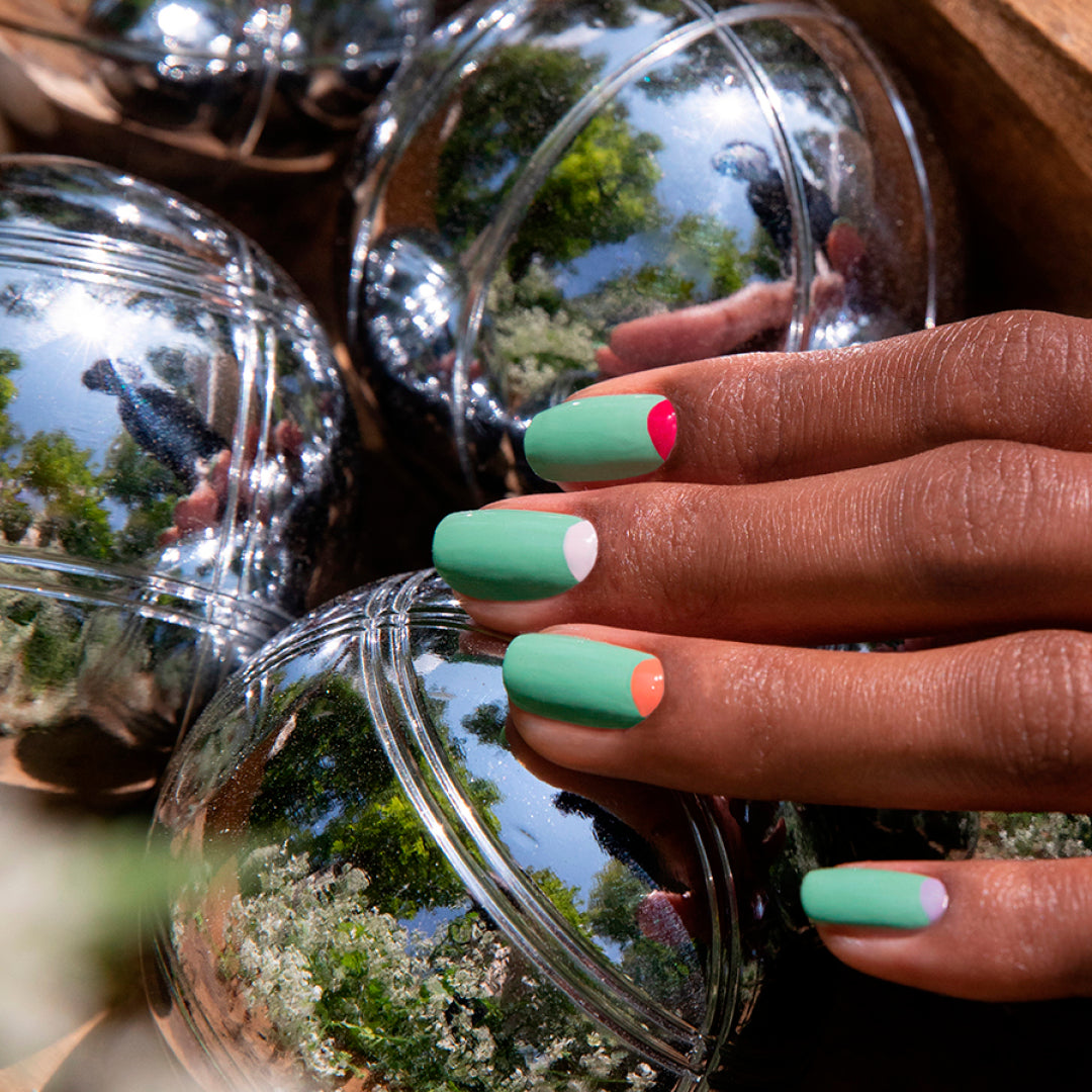A hand with neatly manicured nails painted in Amande by Manucurist rests on shiny metallic balls that reflect the lush greenery.