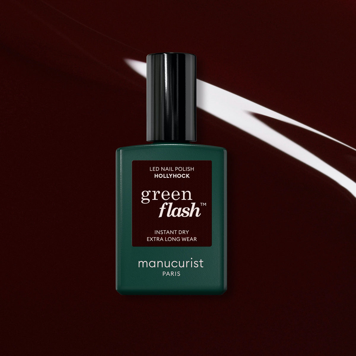 A bottle of Manucurist Hollyhock Green Flash gel polish, a rich deep red shade, displayed on a dark maroon background with a white streak.