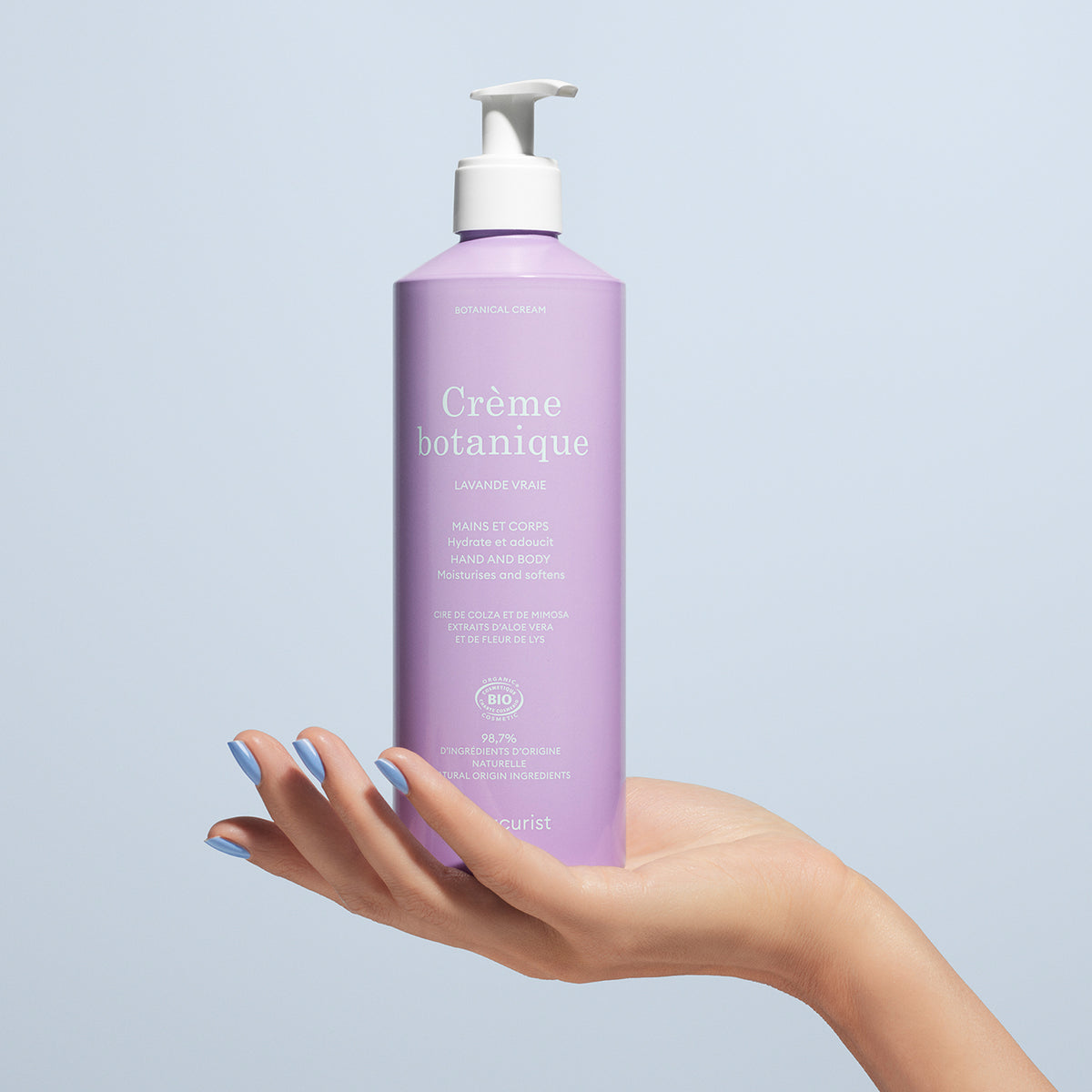 A hand with manicured nails holds a tall lavender pump bottle labeled "Lavande Hand Cream - Cosmos Organic - 500ml" by Manucurist against a plain light blue background.