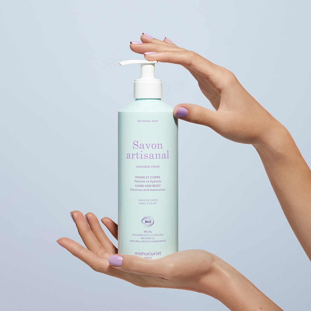 A person with pastel purple nails holds a 500ml Manucurist Lavande Artisan Soap (Cosmos Organic) with a white pump, against a plain light blue background.