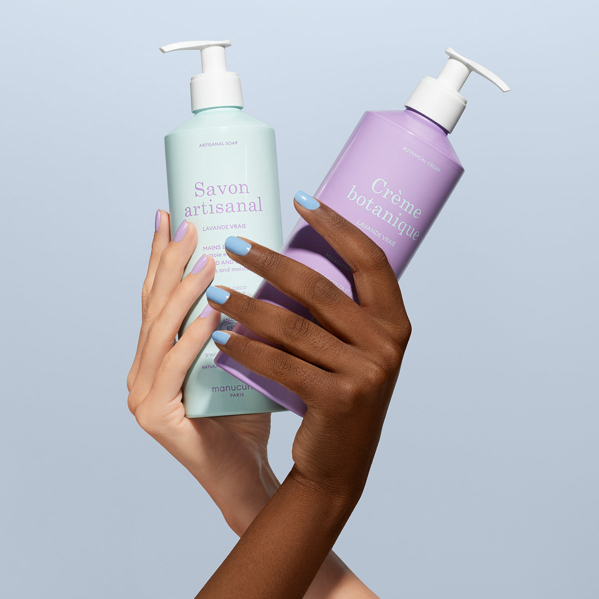 Two hands with manicured nails—one light, one dark—hold a pastel pump bottle of Manucurist Soothing Lavande Hand Body Cream - Cosmos Organic against a light blue background.