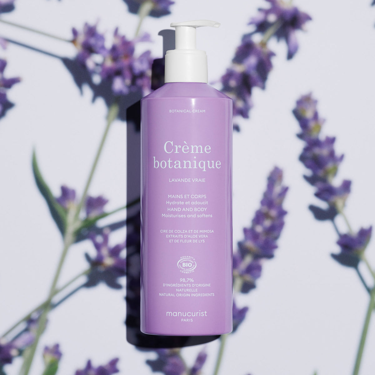 A lavender bottle of Manucurist Lavande Hand Cream - Cosmos Organic (500ml) stands upright, surrounded by blurred lavender sprigs. The label highlights its hydrating, natural, and organic qualities.