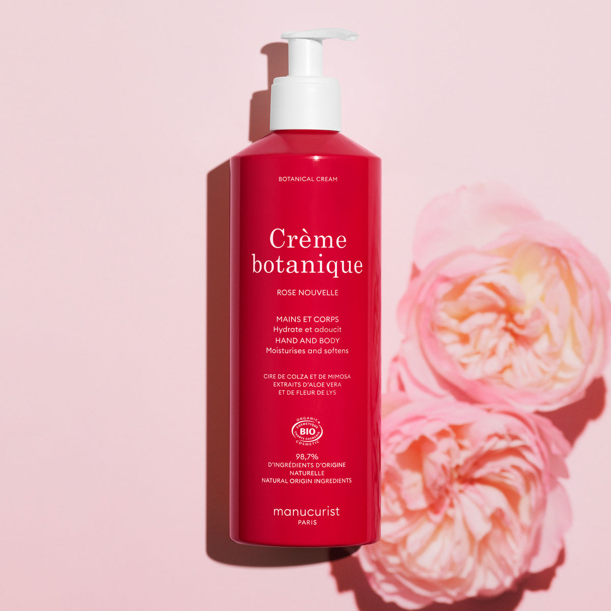 A red bottle of Manucurist Rose Nouvelle Cream - Cosmos Organic (500ml) rests on a light pink surface beside two pink roses. The white label displays detailed text, reflecting its delicate rose scent.