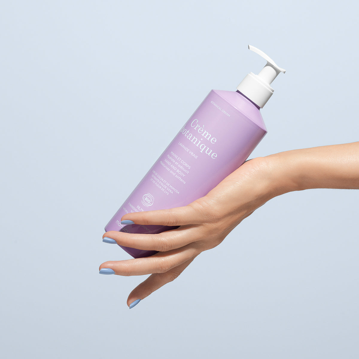A hand with blue-painted nails holds a 500ml bottle of Manucurist Lavande Hand Cream - Cosmos Organic, featuring a white pump dispenser, against a pale blue background.