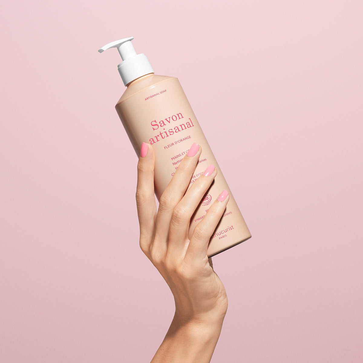 A hand with manicured pink nails holds a beige bottle of Manucurist Fleur d&