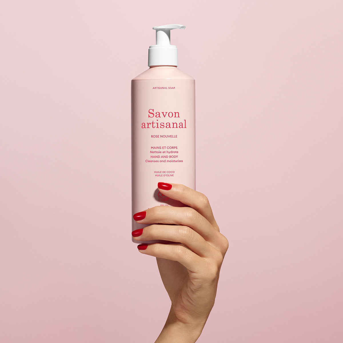 A hand with red nails holds a pink bottle of Manucurist Rose Nouvelle Artisan Soap - Cosmos Organic (500ml), featuring a delicate rose fragrance, set against a light pink background.