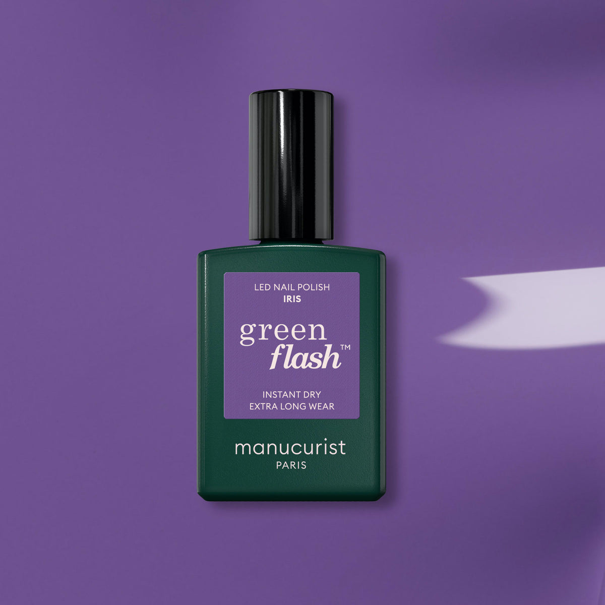 A bottle of Manucurist Iris gel polish rests on a purple background, casting a soft shadow right. The dark green bottle with a purple label highlights this chic shade from the Manucurist brand.