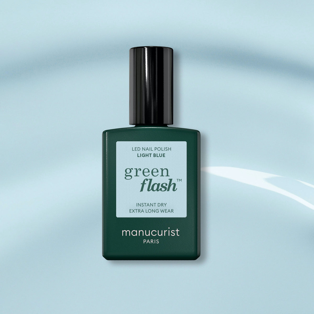 A bottle of Manucurist Light Blue gel polish with a dark green cap sits against a light blue background featuring a subtle glossy reflection.