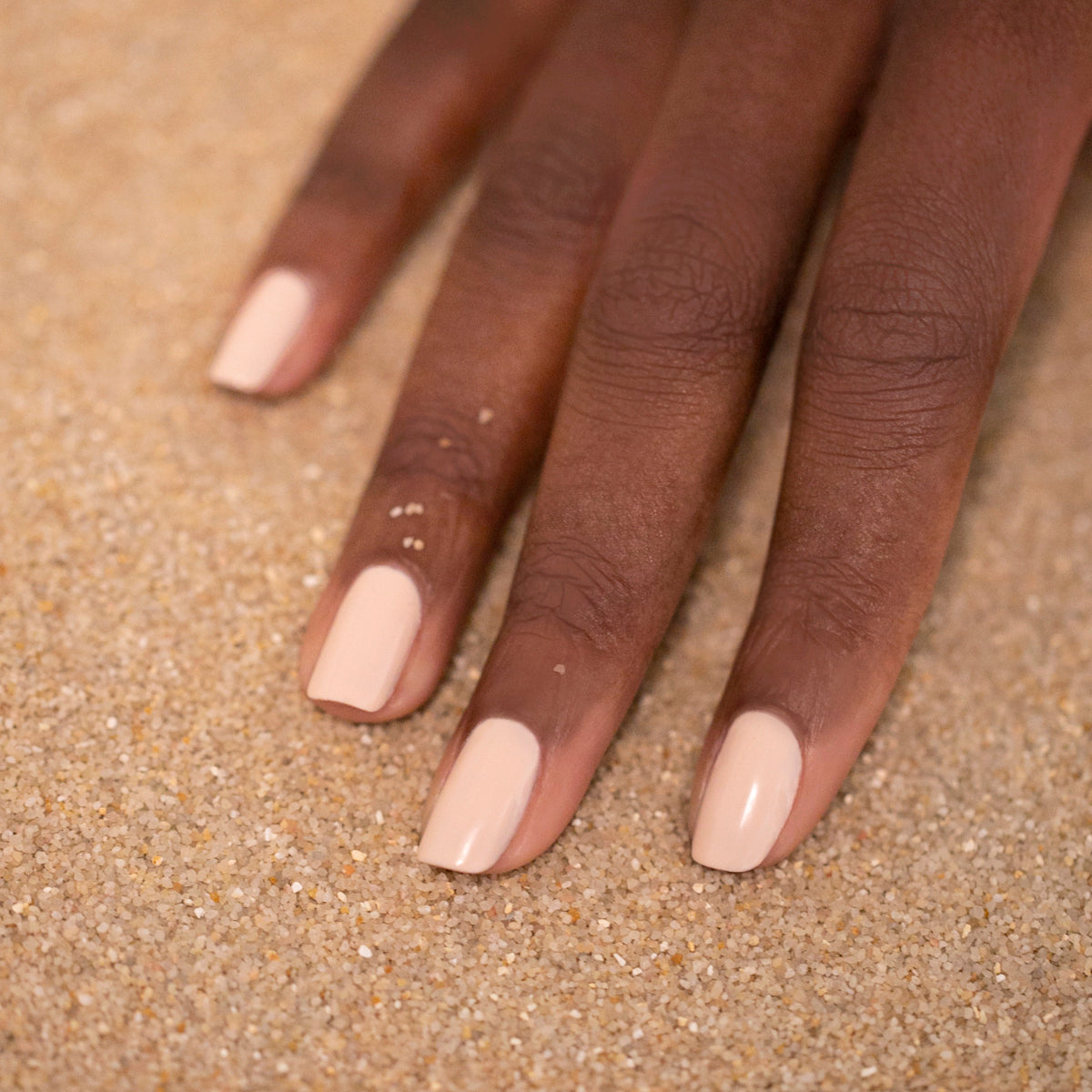 A hand with medium brown skin rests on sand, displaying neatly manicured nails painted in Manucurist&