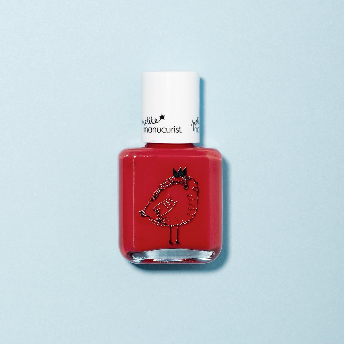 A bottle of Manucurist Lucette the Warbler kids nail polish with a white cap, showing a crowned bird illustration. The red, natural formula is displayed against a light blue background.