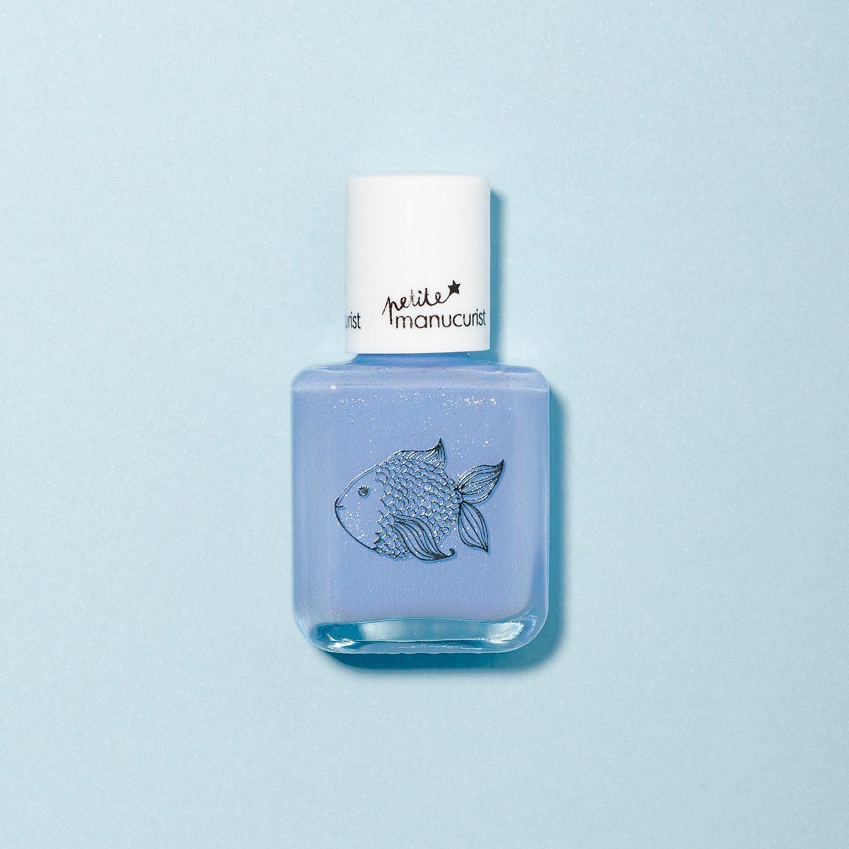 A bottle of Manucurist’s Pomme the Fish kids nail polish in light blue, non-toxic formula with a white cap and a fish illustration on the front. The background is pale blue to match the polish shade.