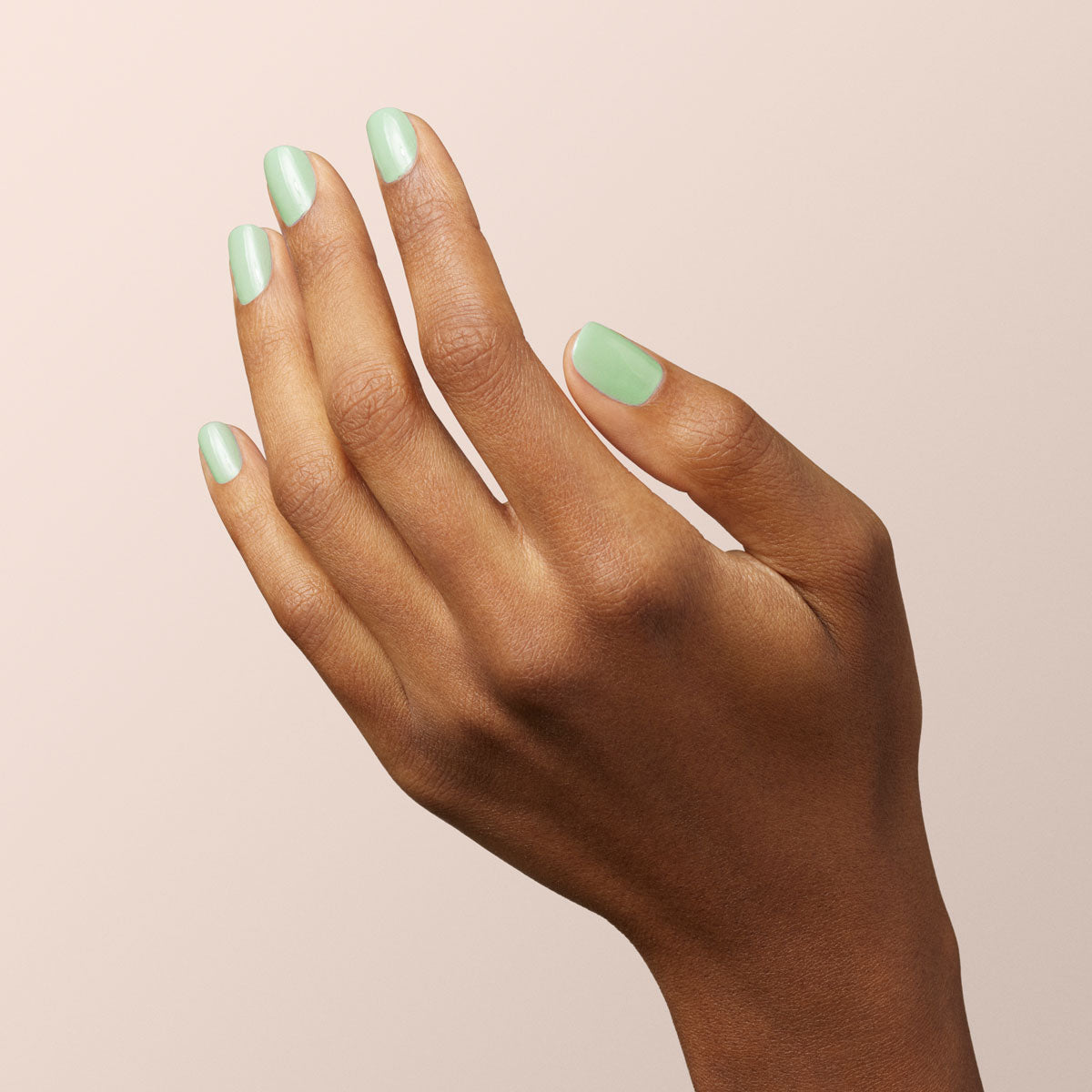 A hand with medium brown skin is shown against a light beige background. The nails are neatly manicured and painted with Mint by Manucurist, a delicate pastel green polish, giving them a glossy, refined touch.