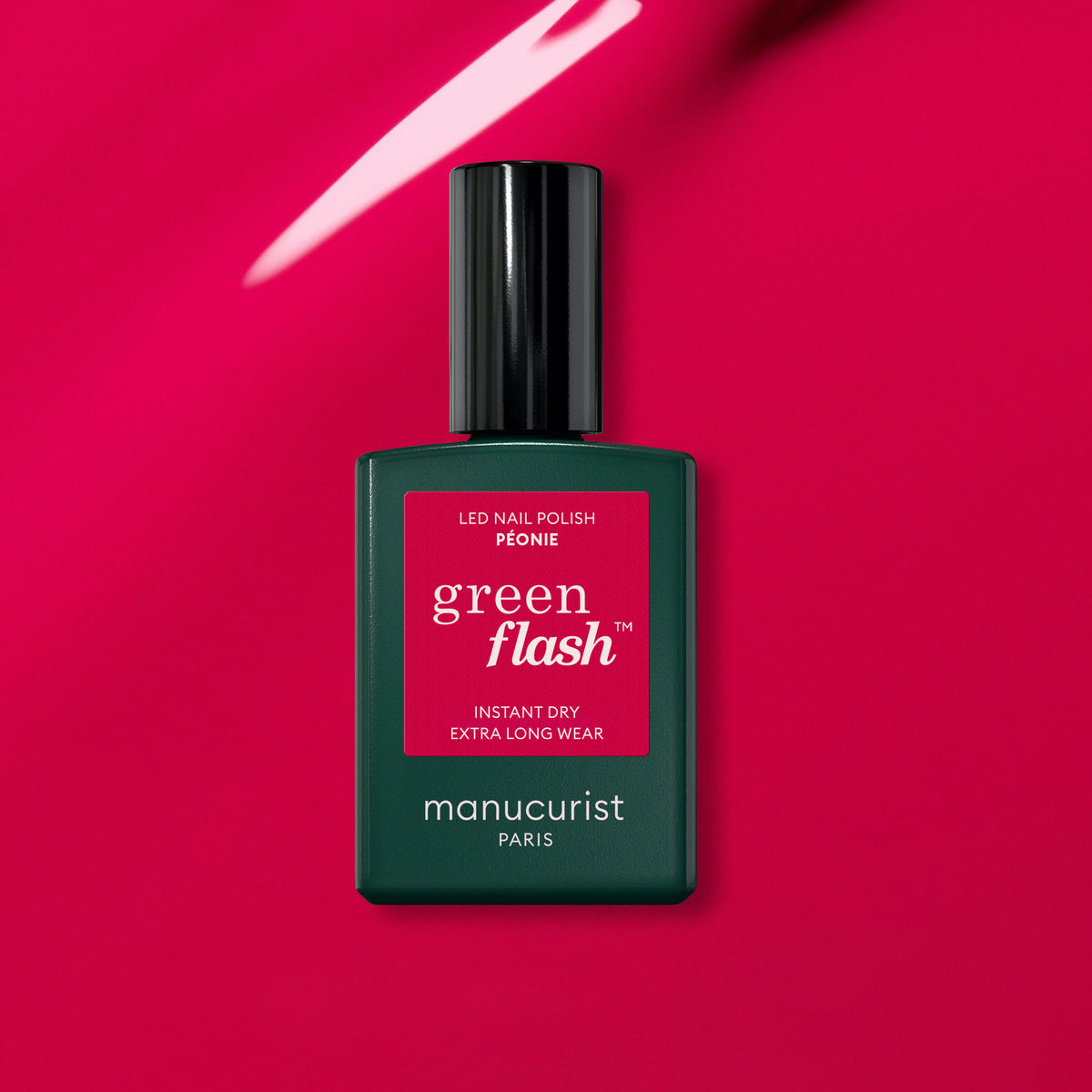 A bottle of Manucurist Péonie Green Flash gel polish stands against a glossy bright pink background, with its green bottle, black cap, and red label showcasing the stunning pink shade.
