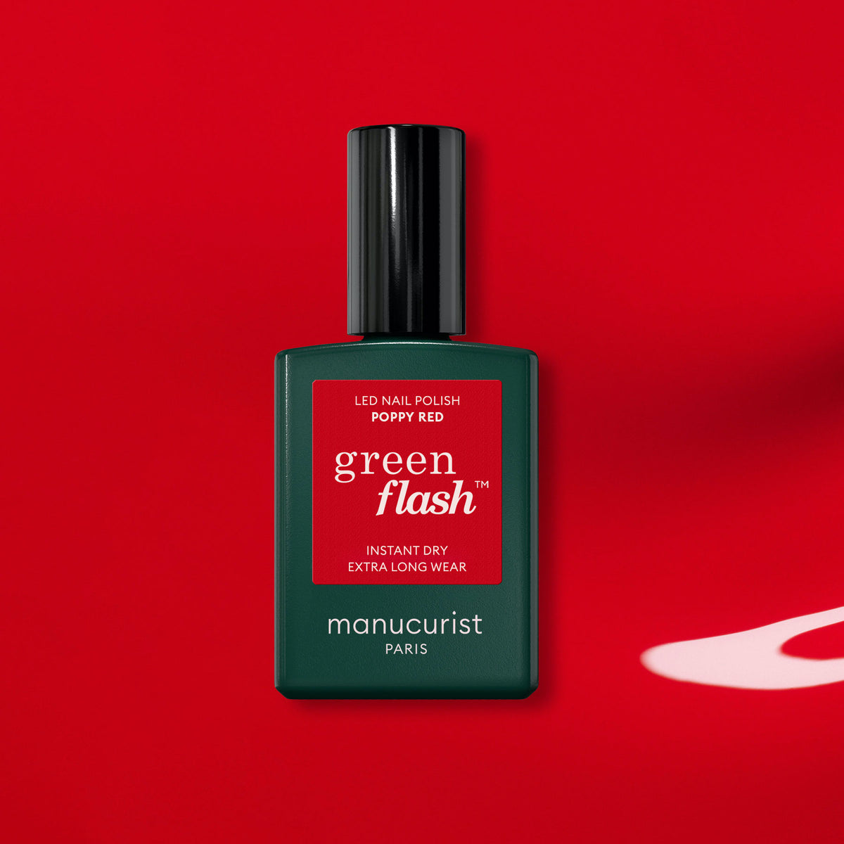 A green bottle of Manucurist-test Poppy Red - Free sits on a vibrant red background with a white shadow, showcasing the orangey red nail polish.
