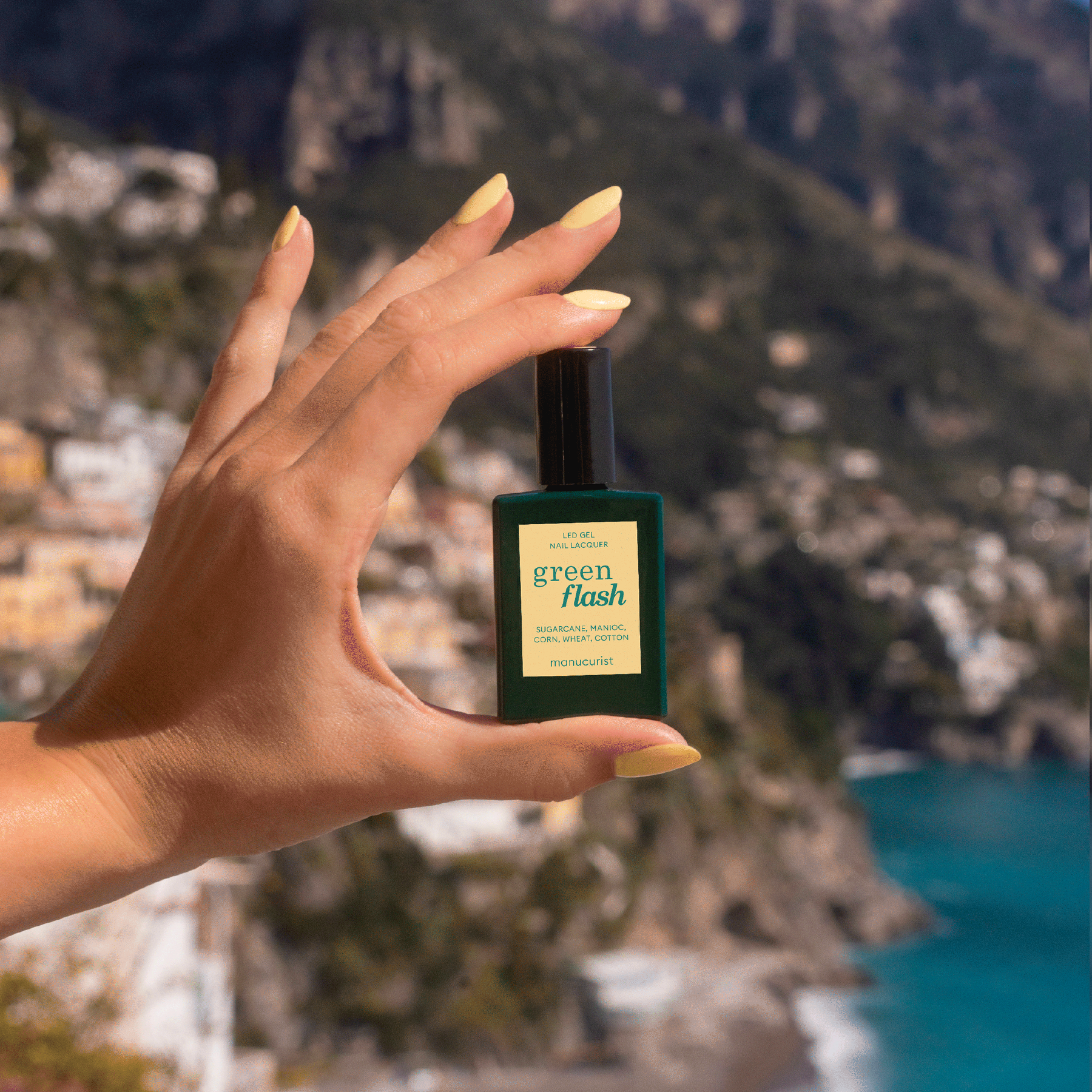 A hand with pastel yellow Manucurist Mimosa gel polish holds a Green Flash bottle against a blurred coastal cliff and sea backdrop.
