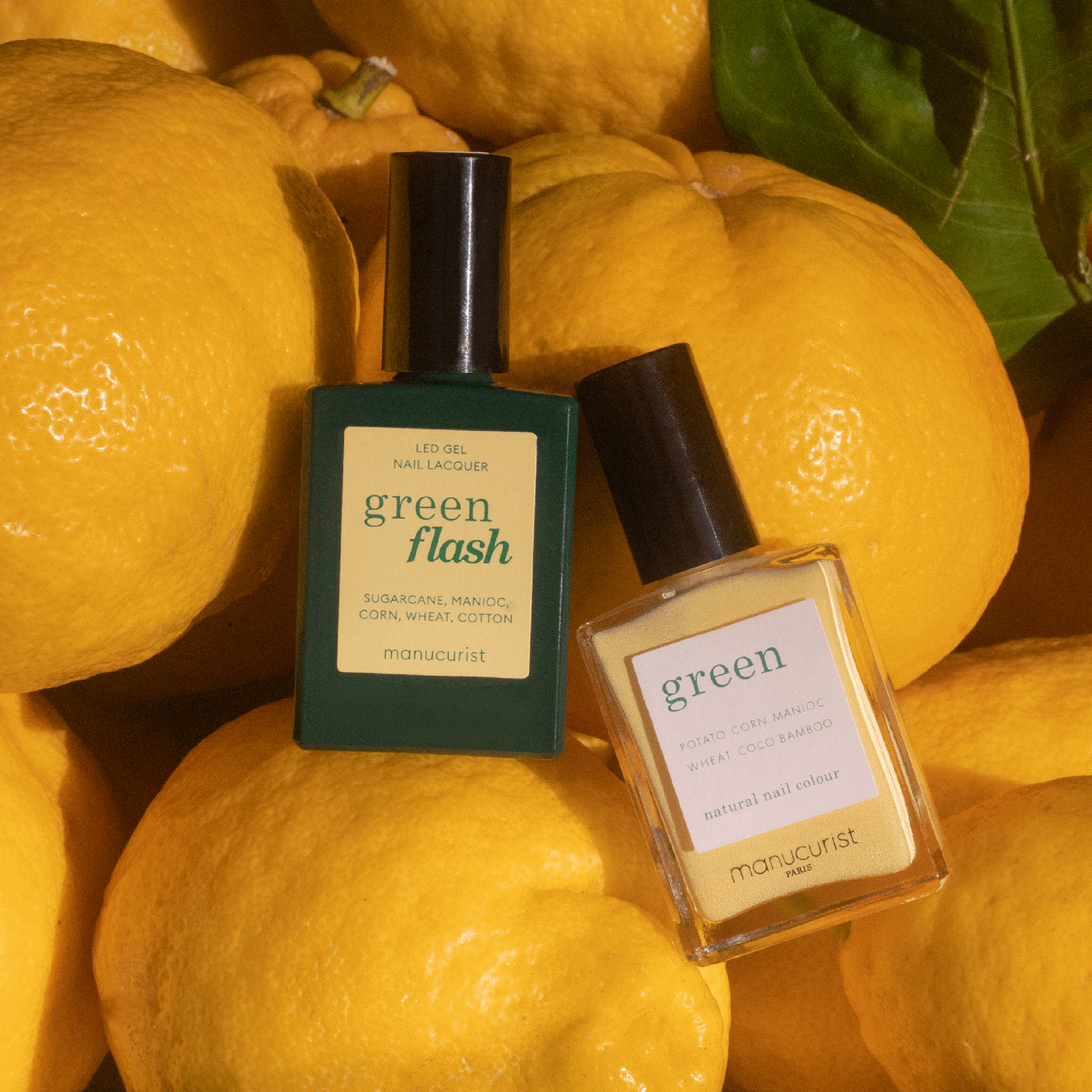 Two bottles of Manucurist nail polish, including the Mimosa shade, are placed on fresh yellow lemons with green leaves. The labels read "green flash" and "Mimosa," complementing the vibrant pastel yellow setting.