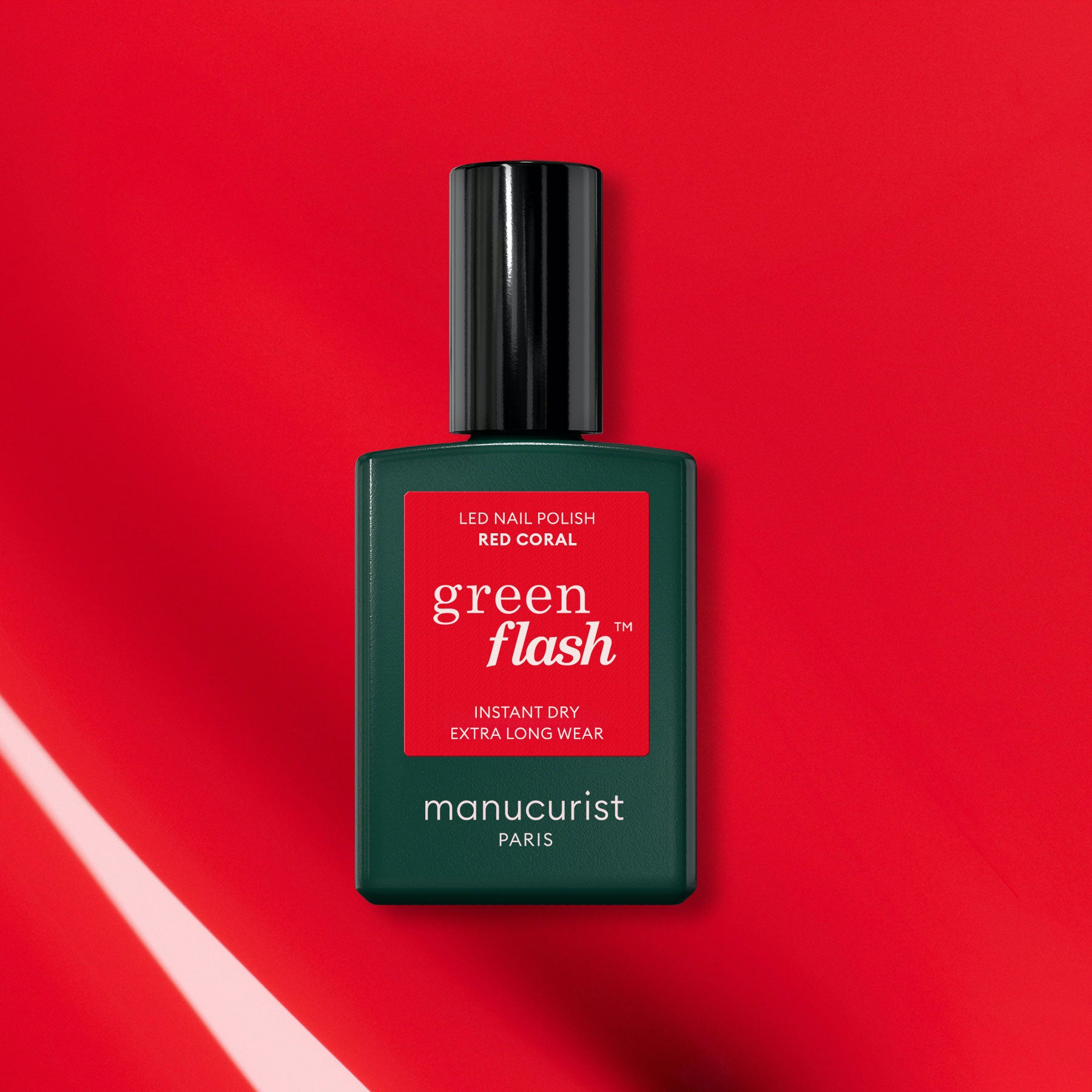 A green bottle of Manucurist-test Red Coral OLD LED nail polish is displayed on a vibrant red background with light reflections, showcasing its long-lasting gel nail polish formula.
