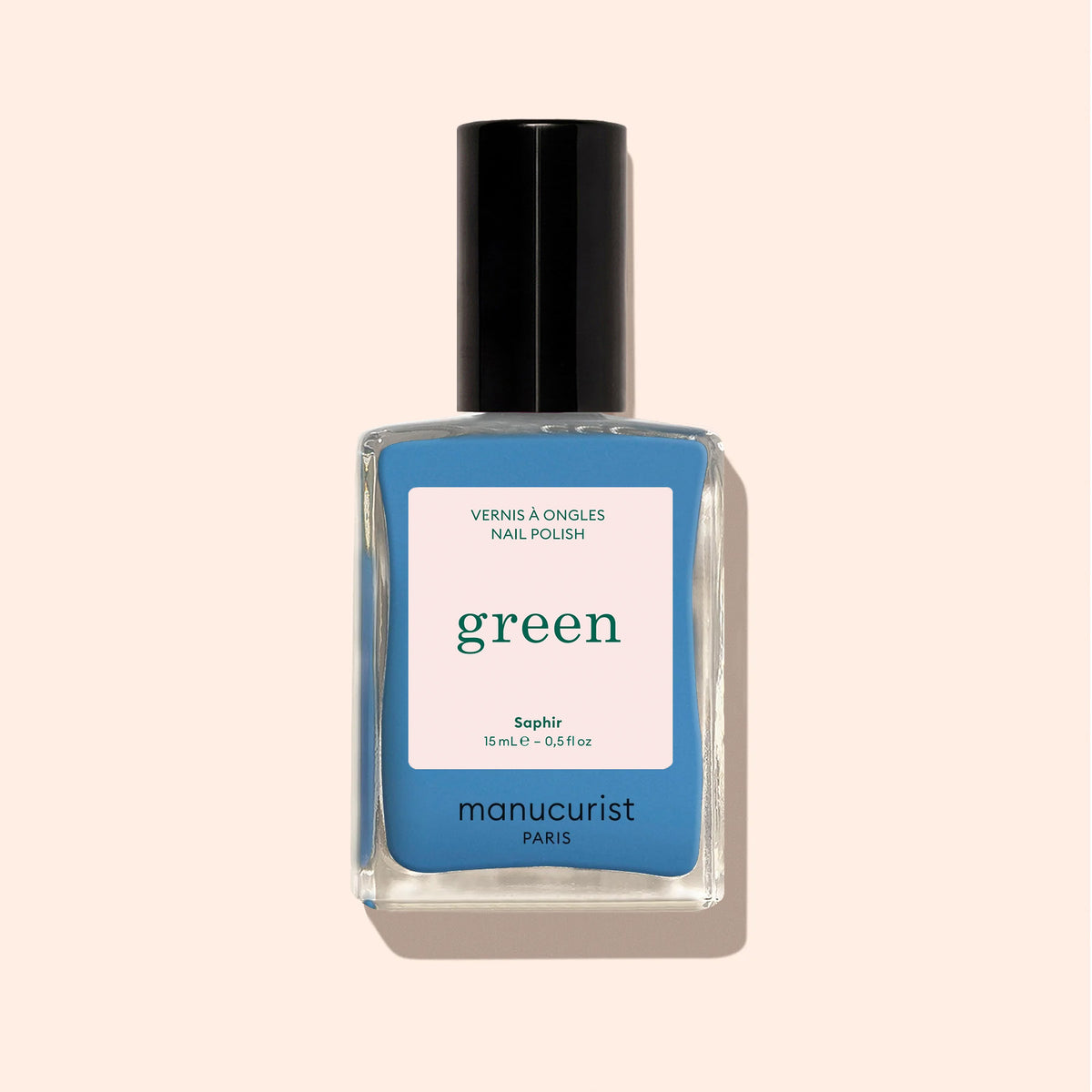Manucurist’s Saphir nail polish, a jewel-toned azure blue with a black cap and beige label, stands out against the light beige background.
