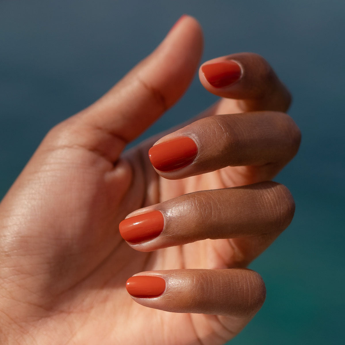 A close-up of a hand with medium brown skin shows neatly manicured nails painted in Manucurist’s Terracotta, a glossy scorched red earth shade—perfect for summer—held against a blurred blue background.