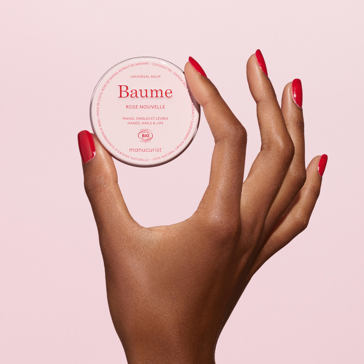 A hand with neatly manicured, red-painted nails holds a round container of Manucurist Natural Nourishing Balm - Cosmos Organic—a multipurpose, natural skincare balm—against a light pink background.