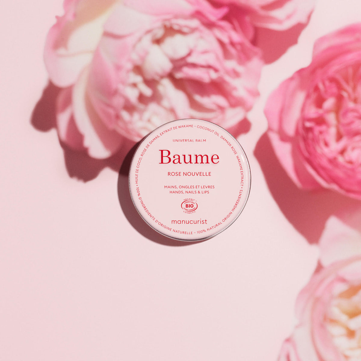 A round container of Manucurist Natural Nourishing Balm - Cosmos Organic rests on a light pink surface, surrounded by pink flowers and soft shadows. The label highlights this multipurpose balm to soothe and repair hands, nails, and lips.