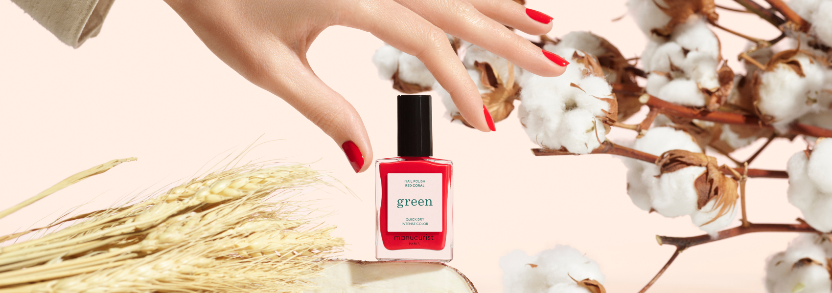 A hand with red-painted nails reaches for a red nail polish bottle labeled green placed among cotton branches and wheat stalks on a light background.