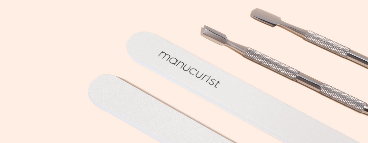A manicure set with two metal cuticle pushers, two white nail files labeled manucurist, and a blue oval tool marked M7XQ on a light beige background.