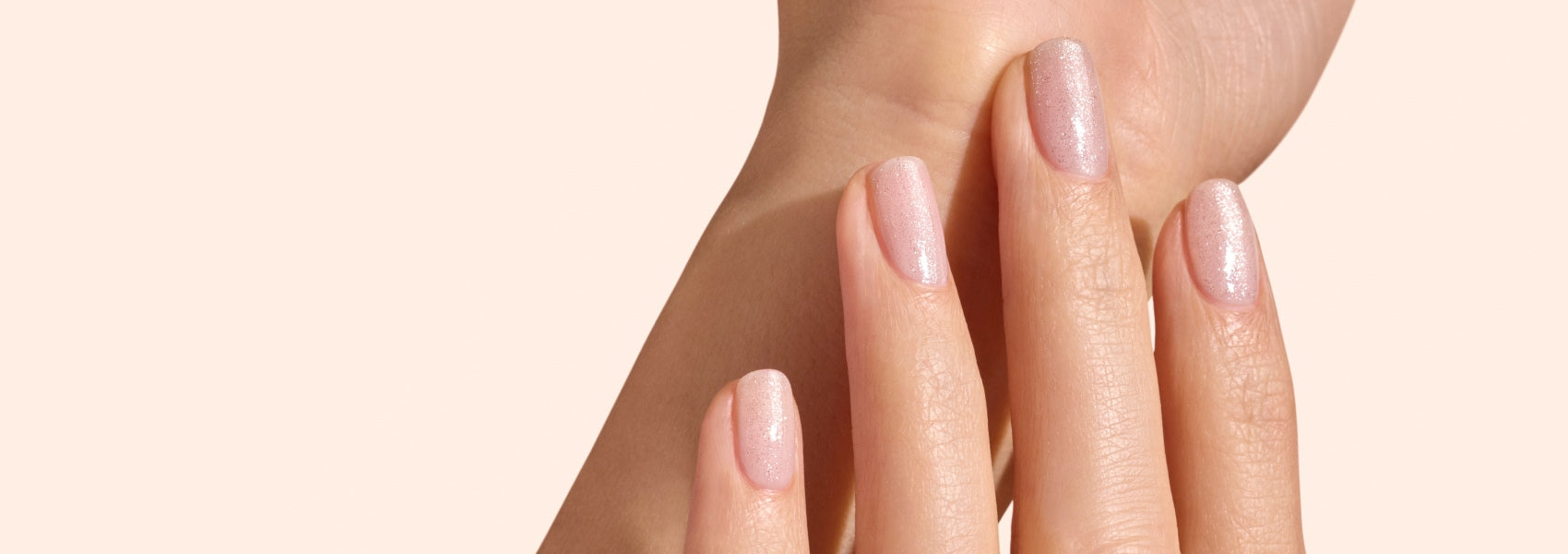 A close-up of a hand with well-manicured nails painted in a light, shimmery nude color, lightly touching the wrist of the other hand against a soft beige background.