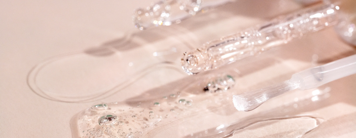 Close-up of glass droppers and a brush applicator with clear, gel-like skincare serum spilled on a light surface, showing bubbles and reflections in the liquid.