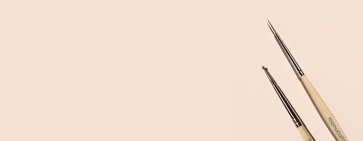 Two fine-tipped nail art brushes with wooden handles positioned on the right side of a pale beige background. The rest of the image is empty space.