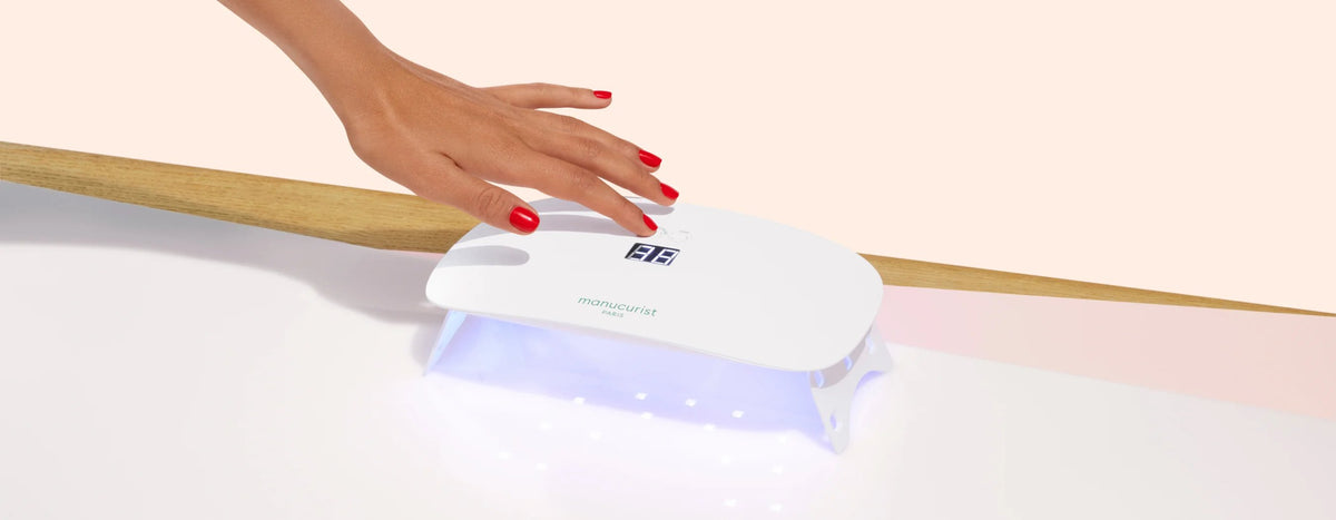 A hand with red nail polish is placed under a white LED nail lamp with digital display, curing gel polish. The scene has a minimalist background with a light wooden accent.