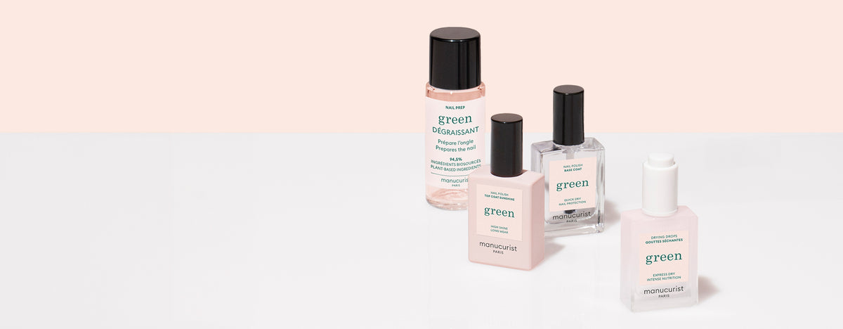 Four manicure products in clear and frosted bottles labeled green by Manucurist are arranged on a white and pale pink background. Products include base, top coat, remover, and nail polish.