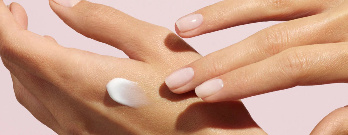 A close-up of a hand applying a dollop of white lotion to the back of another hand, both with neatly manicured nails and a light pink background.