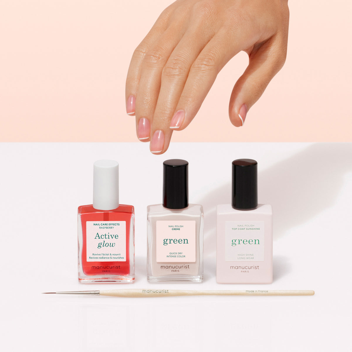 A hand with a glossy manicure hovers above three bottles of Manucurist French Manicure Glow nail polish and a thin nail art brush on a white surface with a pale pink background.