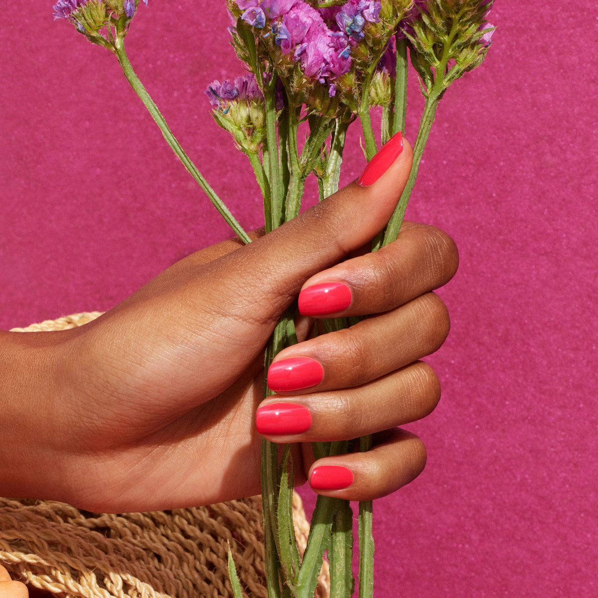 A hand with glossy, reddish pink Manucurist Bougainvillea nail polish holds a bouquet of small purple flowers and bougainvillea against a textured pink background.