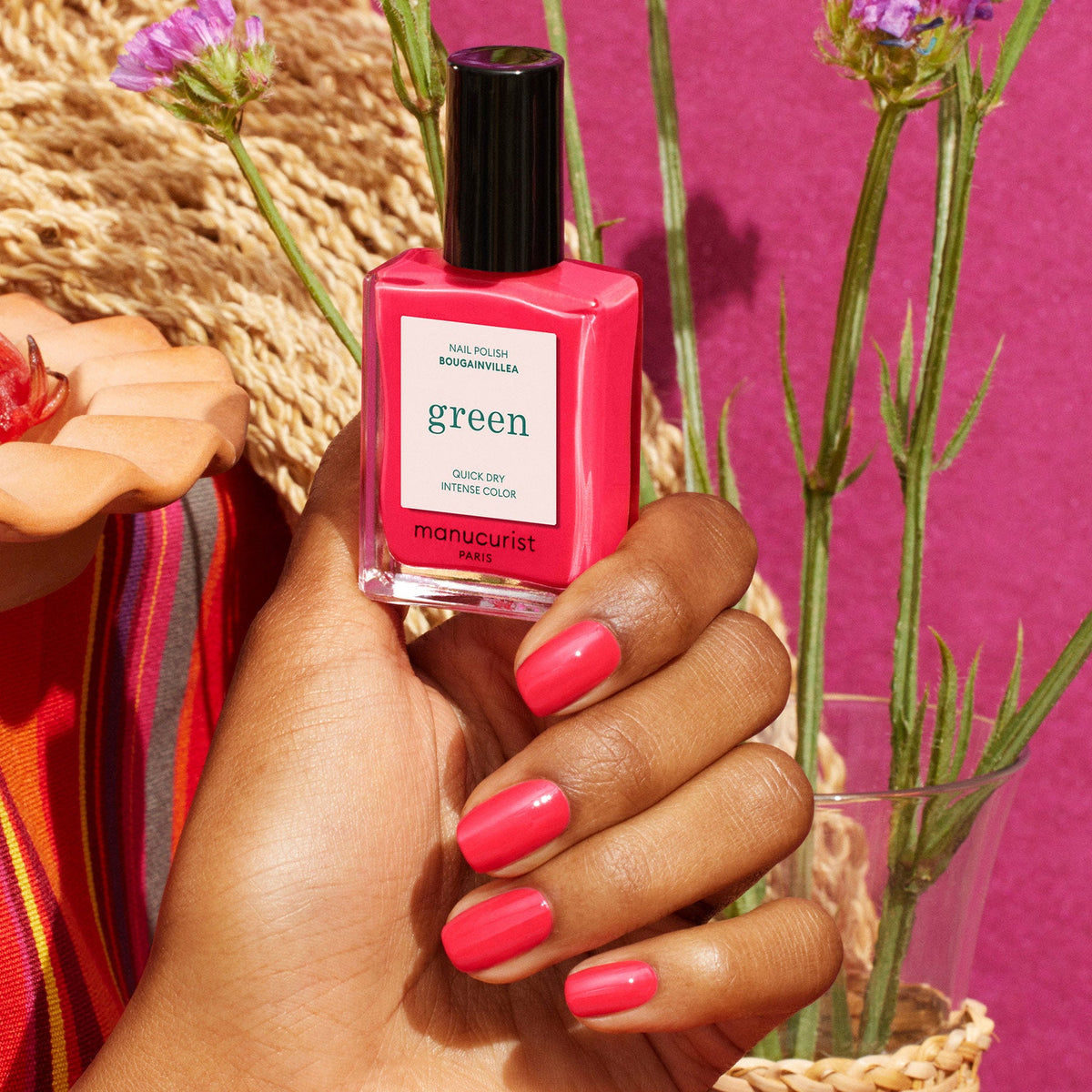 A hand with neatly manicured nails painted in Bougainvillea, a stunning reddish pink polish by Manucurist, holds the bottle. In the background are flowers, a woven hat, and a magenta surface.
