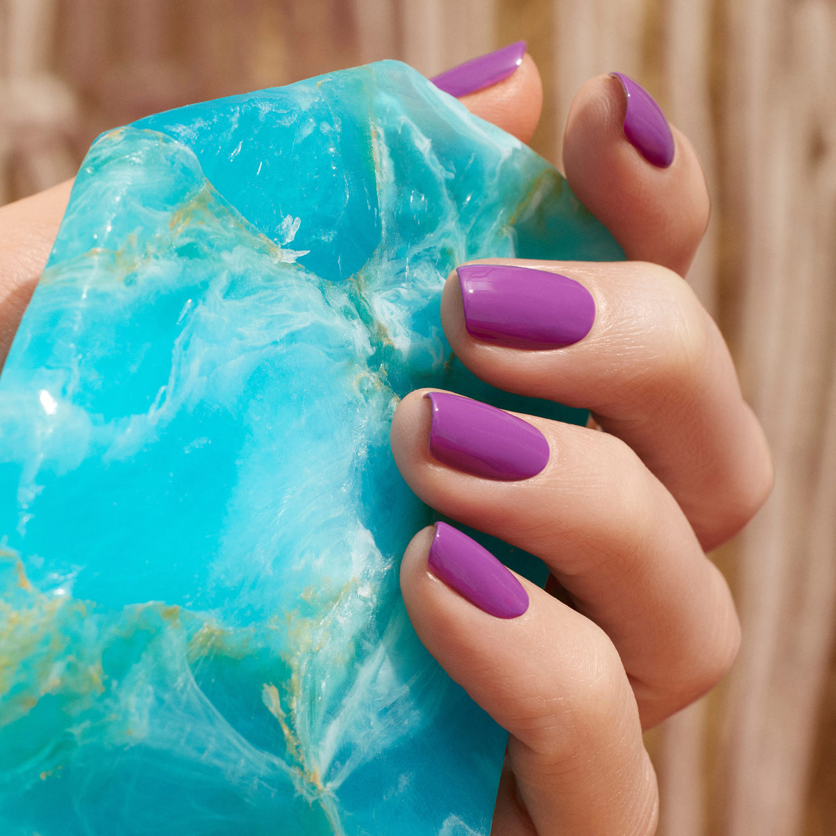 A hand with neatly manicured nails painted in Manucurist’s Violette gel polish holds a large, vivid blue-green gemstone against a blurred wooden background.