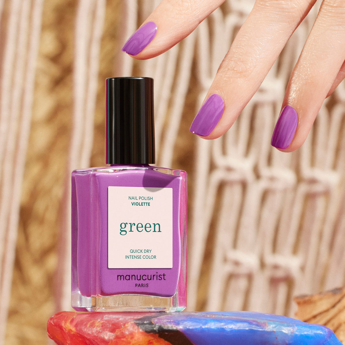 A hand with retro purple-painted nails reaches for a matching bottle of Manucurist Violette nail polish, set on a colorful surface against a blurred woven background.