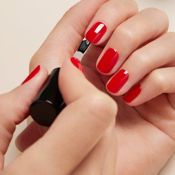 Someone applies Manucurist Sunshine Top Coat, a glossy gel-effect nail polish, to their fingernails with a brush—painting one hand while holding the bottle in the other.
