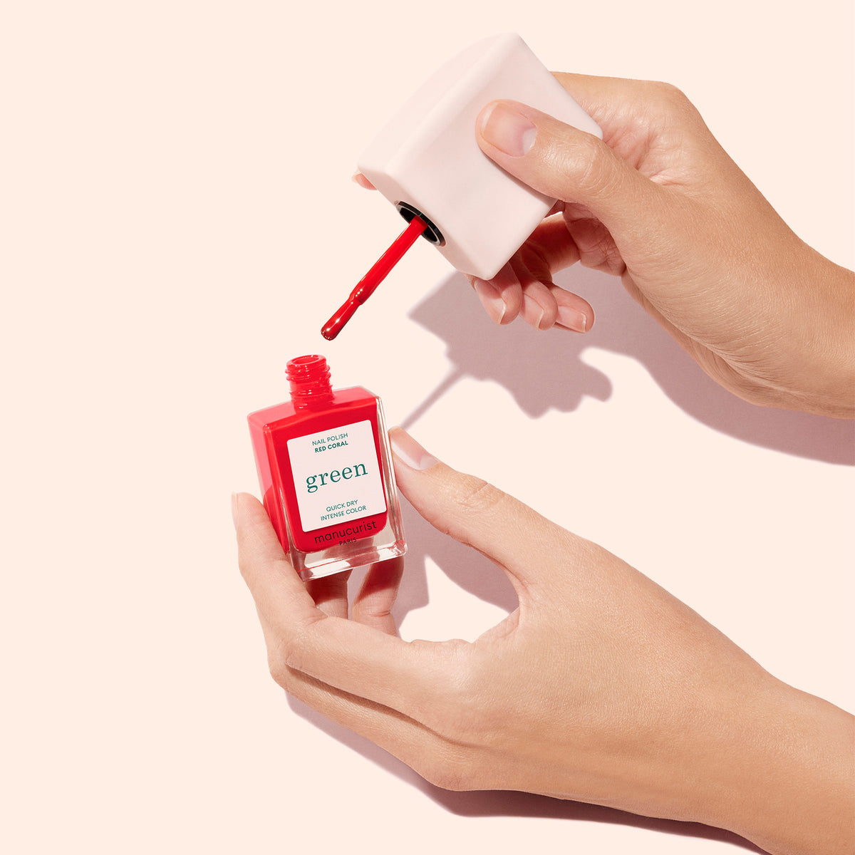 A hand holds an open bottle of Manucurist Mani Helper nail polish in bright red, labeled "green," while another hand holds the white cap with brush applicator, ready for a perfect manicure against a light beige background.