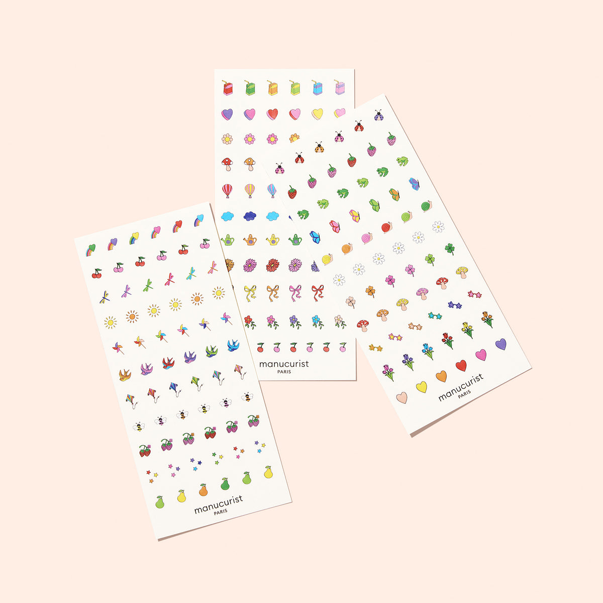 Three Manucurist Nail Art Stickers sheets are displayed on a light pink background, showcasing summer-inspired nature designs—flowers, fruits, hearts, stars, and bows—arranged in neat rows.