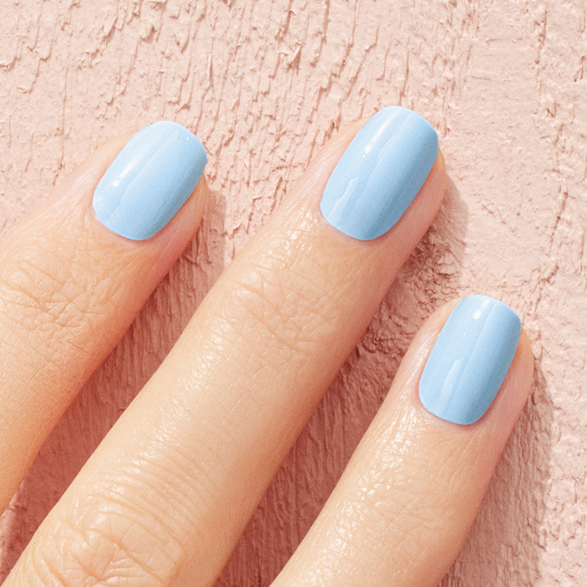 A hand with neatly manicured nails painted in Manucurist’s Bubble, a light blue polish with subtle grey undertones, is shown against a textured pink background.